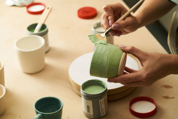 Close-up of girl painting clay mug with glaze. Woman coloring pottery in workshop with a paintbrush. Painter in green apron glazing clay pot.