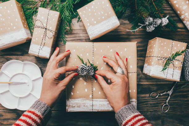 Woman´s hands wrapping Christmas presents on brown paper decorated with painted snow, fir branches and pinecones on a rustic wooden board. Top view
